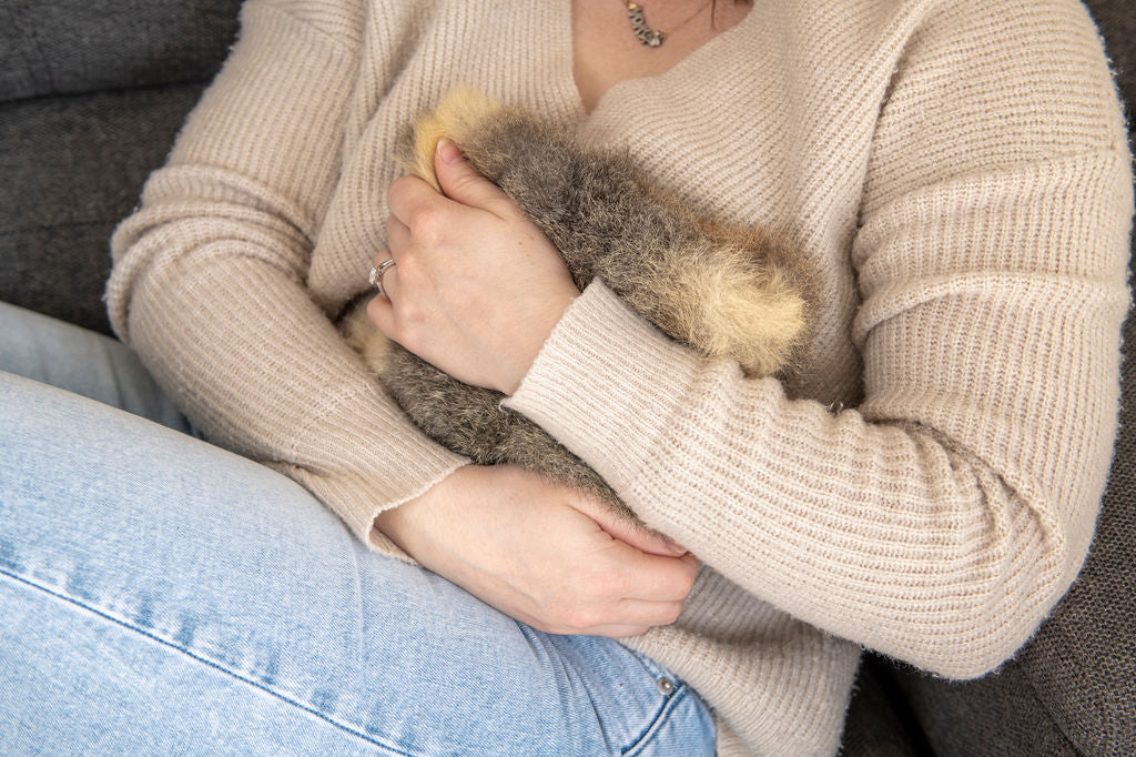 Person holding a small fluffy wheat bag cover in their arms, wearing a beige sweater and blue jeans.